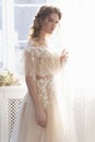 Beautiful natural redhead girl bride, with nude makeup, wearing a white dress, stands at the window next to a transparent curtain Royalty Free Stock Photo