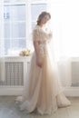 Beautiful natural redhead girl bride, with nude makeup, wearing a white dress, stands at the window next to a transparent curtain Royalty Free Stock Photo