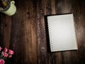 A beautiful notebook is placed on desk dark brown wood with a pink rose in the lower left corner. And a leaf in the top left corne