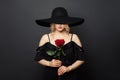Beautiful passionate woman wearing wide black broad brim hat with red rose flower on black background Royalty Free Stock Photo