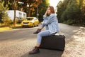 Beautiful pensive girl sits on her suitcase on middle road, waiting a transportation, in sunset background. Royalty Free Stock Photo