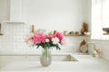 Beautiful peonies in vase on granite countertop on background of stylish white kitchen with wooden shelves and appliances in new Royalty Free Stock Photo