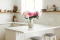 Beautiful peonies in vase on granite countertop island on background of stylish white kitchen with wooden shelves and appliances Royalty Free Stock Photo