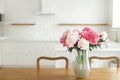 Beautiful peonies in vase on wooden table on background of stylish white kitchen with island, wooden shelves and appliances in new Royalty Free Stock Photo