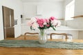 Beautiful peonies in vase on wooden table on background of stylish white kitchen with island, wooden shelves and appliances in new Royalty Free Stock Photo