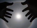 Beautiful photograph of the sun with a circular rainbow surrounded by a bright sky and white clouds with shadows of hands reaching Royalty Free Stock Photo