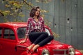 Beautiful pin-up girl in jeans and a plaid shirt posing, sitting on the hood of a red retro car. Royalty Free Stock Photo