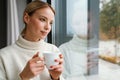 Beautiful pleased woman drinking coffee while leaning on window glass Royalty Free Stock Photo
