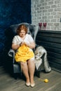 A beautiful plump redhead young woman with freckles sits in an armchair and knits a yellow sweater