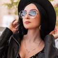 Beautiful portrait chic young woman with sexy lips in fashionable hat in stylish sunglasses in trendy leather jacket outdoors on