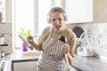 Beautiful positive woman in earphones listening to music and singing into ladle as microphone while cooking in the kitchen. Relax Royalty Free Stock Photo