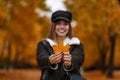 Beautiful positive young woman with a beautiful smile in a trendy hat with a visor in a jacket holds a yellow maple leaf in hands Royalty Free Stock Photo