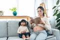 A beautiful pregnant Asian single mother is reading a book comfortably on her living room sofa. With a cute little daughter Royalty Free Stock Photo