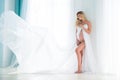 Beautiful pregnant blonde woman stands near the window. Flying effect of white chiffon fabric