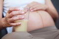 Beautiful pregnant woman drinking a glass of milk and keeping a hand on her bare tummy while sitting