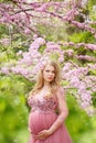 Beautiful pregnant woman in gently pink dress and touching belly is standing near the cherry blossom