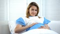 Beautiful pregnant woman holding tummy and smiling at camera, prenatal care