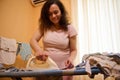 Beautiful pregnant woman ironing baby clothes using electric steam iron, smiling, enjoying her maternity leave. Royalty Free Stock Photo