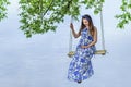 Beautiful pregnant woman mother to be in wreath and blue dress sitting on swing over scenic river in sunset time in summer. Royalty Free Stock Photo