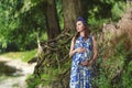 Beautiful pregnant woman mother to be in wreath and blue dress standing in front of scenic roots of tree in sunset time in summer Royalty Free Stock Photo