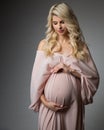 Beautiful Pregnant Woman Portrait in Pink Chiffon Dress over Gray. Mother looking at Belly in Pregnancy Bridal Fashion Gown Royalty Free Stock Photo