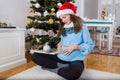 Beautiful pregnant woman with Santa hat sitting on floor and making heart shape gesture with hands on her belly. Christmas tree in Royalty Free Stock Photo