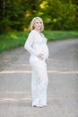 Beautiful pregnant woman in sheer long lacy white maternity dress looking dreamy on lonely road in forest Royalty Free Stock Photo