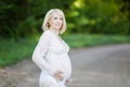 Beautiful pregnant woman in sheer long lacy white maternity dress looking dreamy on lonely road in forest Royalty Free Stock Photo