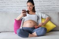 Beautiful pregnant young woman using her mobile phone while eating strawberries on sofa at home Royalty Free Stock Photo