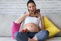 Beautiful pregnant young woman using her mobile phone while eating strawberries on sofa at home Royalty Free Stock Photo