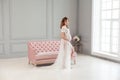 Beautiful pregnant young woman in white dress peignoir standing near pink sofa, holding with love her belly Royalty Free Stock Photo