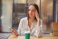 Beautiful puzzled woman wearing bluse sitting in cafe using mobile phone using mobile phone lookign at camera having unhappy