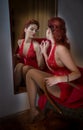 Beautiful red hair girl with long red lace dress posing in front of a large wall mirror. Young attractive redhead, side view