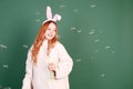 A beautiful red-haired girl with rabbit ears blows soap bubbles on a green background. Easter holidays