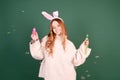 A beautiful red-haired girl with rabbit ears blows soap bubbles on a green background. Easter holidays