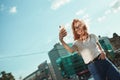 Beautiful red-haired girl stands in the street on the background of the city and takes a selfie on her mobile phone. The girl is