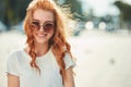 A beautiful red-haired girl in a white T-shirt and sunglasses walks down the street and smiles in frame