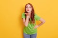 Beautiful red haired lady blowing air kiss to camera isolated over yellow background, looks satisfied, wearing green t shirt and