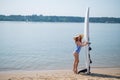 A beautiful red-haired woman in a straw hat, sunglasses and a striped one-piece swimsuit stands on the beach and holds a Royalty Free Stock Photo