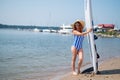 A beautiful red-haired woman in a straw hat, sunglasses and a striped one-piece swimsuit stands on the beach and holds a