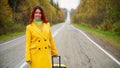 Beautiful red-haired woman in a yellow coat walks with a yellow suitcase along the highway in autumn. Royalty Free Stock Photo