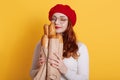 Beautiful red haired young woman holding paper bag with bread on yellow background, satisfied girl smells fresh fragrant long loaf Royalty Free Stock Photo