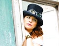 Beautiful redhair steampunk girl with goggles on black hat outside toilet background. Old-fashioned Royalty Free Stock Photo