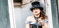 Beautiful redhair steampunk girl with goggles on black hat outside toilet background. Old-fashioned Royalty Free Stock Photo