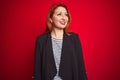 Beautiful redhead business woman wearing elegant jacket over isolated red background looking away to side with smile on face, Royalty Free Stock Photo