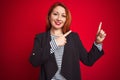 Beautiful redhead business woman wearing elegant jacket over isolated red background smiling and looking at the camera pointing Royalty Free Stock Photo