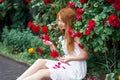 Portrait of a pretty redhead girl dressed in a white light dress on a background of blooming roses. Outdoor Royalty Free Stock Photo