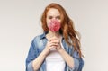 Beautiful redhead girl eating big colorful lollipop, light studio background Royalty Free Stock Photo