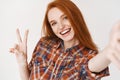 Beautiful redhead girl smiling happy and showing peace sign while taking selfie on smartphone, front camera view, white Royalty Free Stock Photo