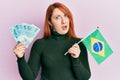 Beautiful redhead woman holding 100 brazilian real banknotes and brazil flag in shock face, looking skeptical and sarcastic,